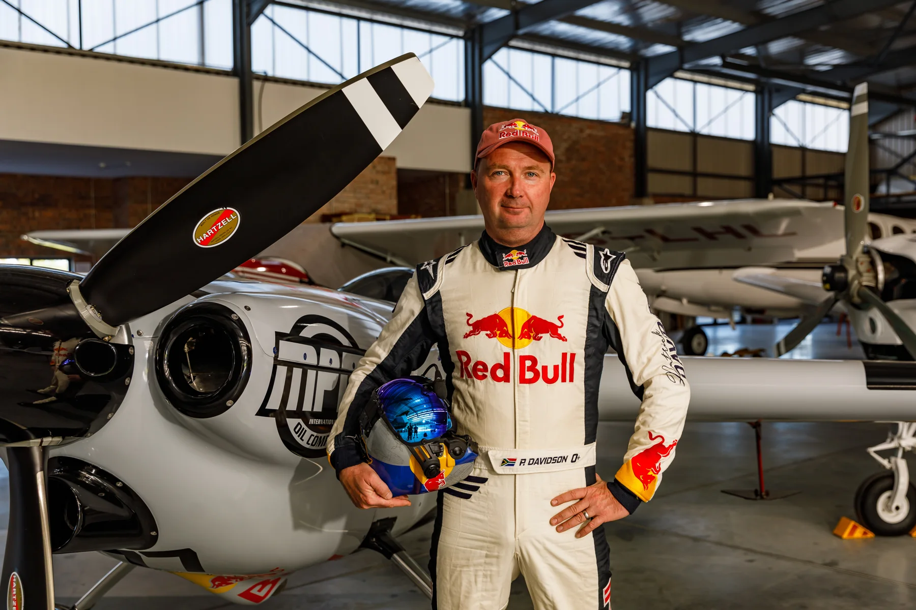 Red Bull pilot Patrick Davidson in flying suit and cap, holding his helmet beside the race aircraft in a hangar.